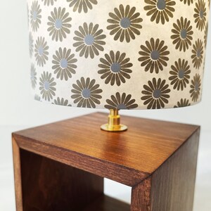 May include: A small wooden table with a shelf and a lamp with a white lampshade featuring a brown and grey floral pattern.