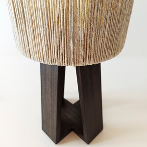 Może przedstawiać: Nowoczesna lampa z cylindrycznym kloszem owiniętym naturalną liną. Klosz spoczywa na ciemnobrązowej, geometrycznej drewnianej podstawie. Lampa emituje ciepłą poświatę od wewnątrz, tworząc przytulną atmosferę. Projekt jest minimalistyczny i stylowy.