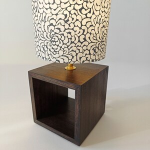 May include: A dark brown wooden lamp base with a square open shelf and a white lampshade with a black floral pattern.