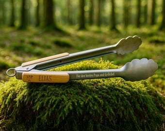May include: A pair of stainless steel and wood grilling tongs with the name "ELIAS" engraved on the wooden handle. The tongs are resting on a moss-covered surface, with a forest background.
