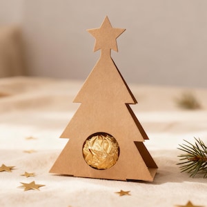 Puede incluir: Una caja de regalo marrón con forma de árbol de Navidad de cartón con una estrella en la parte superior. La caja contiene un chocolate envuelto en oro. Confeti dorado en forma de estrella y una ramita de pino están esparcidos sobre una superficie beige.