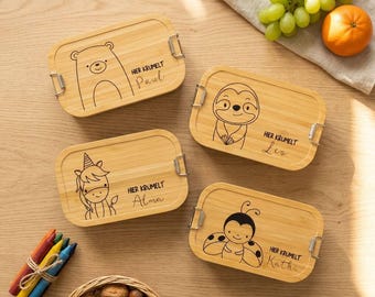 May include: Four rectangular bamboo lunch boxes with metal latches, each featuring a black line drawing of an animal and a name. The designs include a bear labeled Paul, a sloth labeled Leo, a unicorn labeled Alma, and a ladybug labeled Kathi.