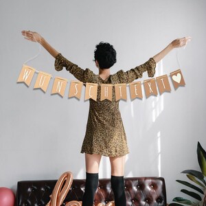 May include: A brown paper banner with the text "WUNSCHTEXT" in white lettering and a small white heart on the right side. The banner is strung up with white string and held by a person with their arms outstretched.