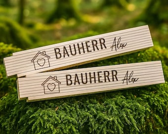 May include: Two wooden signs with the text "Bauherr Alex" and a house icon with a heart inside. The signs are on a bed of green moss.