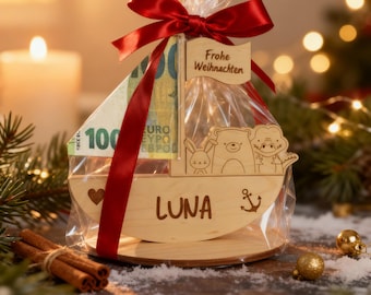 Puede incluir: Una caja de regalo de madera en forma de barco con un lazo rojo, que contiene dinero y una pequeña bandera de madera que dice "Frohe Weihnachten". El barco tiene el nombre "LUNA" y una pequeña ancla grabada. El barco también tiene una pequeña ilustración de un oso, un conejo y un personaje.