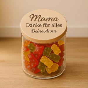 May include: A clear glass jar filled with colorful gummy bears. The jar has a wooden lid with the words "Mama Danke für alles Deine Anna" engraved on it. The gummy bears are red, yellow, and green.