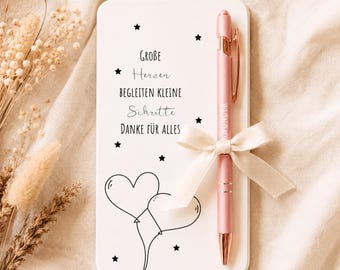 May include: A white rectangular notepad featuring black star icons, a heart-shaped balloon illustration, and the German text "Große Herzen begleiten kleine Schritte Danke für alles." A pink pen with rose gold accents is attached with a cream ribbon.