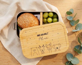 May include: A rectangular metal lunch box with a bamboo lid featuring the engraved text "HIER KRÜMELT Alma" and illustrations of a bear, deer, llama, and koala. The open box contains a round bread roll and a small portion of green grapes.