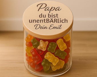 May include: A clear glass jar filled with colorful gummy bears. The jar has a wooden lid with the words "Papa du bist unentBÄRLich Dein Emil" engraved on it. The gummy bears are in various colors, including red, orange, yellow, and green.