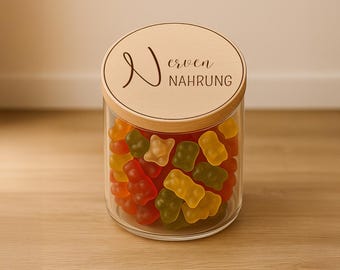 May include: A clear glass jar filled with colorful gummy bears. The jar has a wooden lid with the words "Nerven Nahrung" engraved on it. The gummy bears are various colors, including red, yellow, orange, and green.