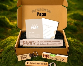 May include: A brown cardboard gift box with the text "Für den besten Papa" on the lid. Inside are cards, a ruler, a keychain, and a bottle opener, all with the theme of "Dad". The box is on a mossy surface.