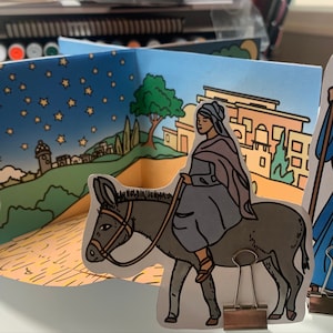 Peut inclure: Une scène d'artisanat en papier représentant le voyage des trois rois mages. La scène comprend un âne, un homme monté sur un âne et un homme avec un bâton. L'arrière-plan présente un ciel nocturne étoilé, une ville et une colline.