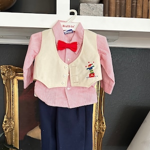 May include: A children's formal outfit, including a red and white striped button-down shirt, a cream-colored vest with an embroidered design, a red bow tie, and navy blue pants. The outfit is hanging on a white hanger.