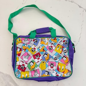May include: A purple shoulder bag featuring a colorful pattern of Looney Tunes characters. The bag has a green shoulder strap and handles, with a white dotted background. The bag is rectangular and has a zipper closure.
