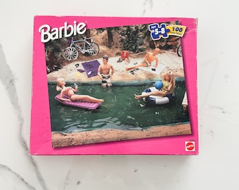May include: A pink Barbie 100 piece jigsaw puzzle box featuring a pool scene with four dolls, a bicycle, and a blue inflatable ring. The box has the text "Barbie" and "Ages 5-8" on the front.
