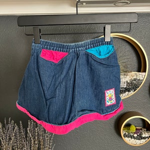 Puede incluir: Una falda de mezclilla azul oscuro con cintura elástica, que presenta un bolsillo rosa brillante a la izquierda y un bolsillo turquesa a la derecha. El dobladillo está adornado con un borde de tela rosa ancho y un fino acento turquesa. Un pequeño parche rectangular muestra un personaje de dibujos animados.