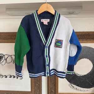 May include: A blue, white, and green knitted cardigan with a patch on the front that says "GIPPSLAND". The cardigan has a button-up front and a V-neck.