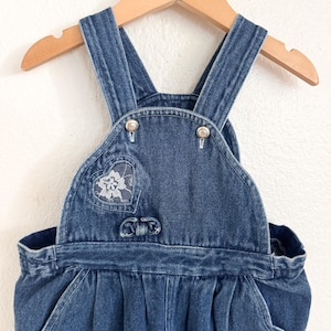 Puede incluir: Vestido de peto de mezclilla azul con tirantes ajustables, un parche en forma de corazón con un diseño floral y botones decorativos. El vestido está colgado en una percha de madera.