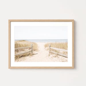 May include: A framed wall art print featuring a sandy path leading to the ocean, flanked by wooden fences and tall, dry beach grass. The scene is captured in soft, neutral tones with a light oak-colored frame and a white mat border.