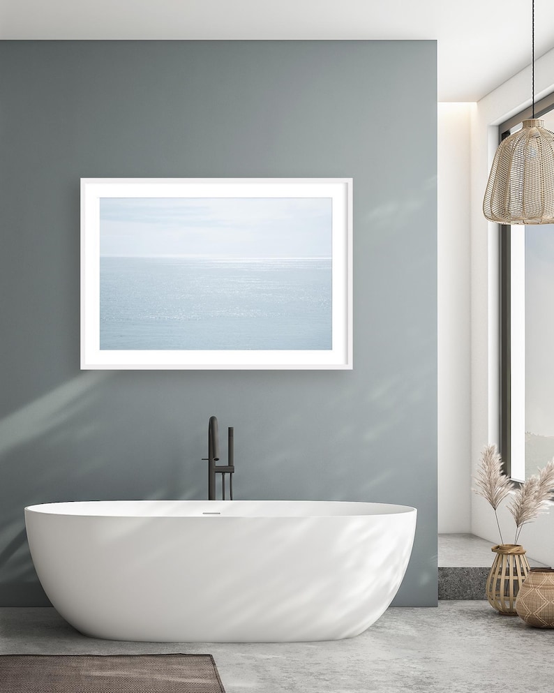 May include: A modern bathroom with a white oval bathtub, a framed seascape print, and a black faucet. The walls are painted a muted teal, and the floor is concrete. A woven pendant light hangs from the ceiling, and decorative vases add a natural touch.
