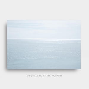 May include: A serene seascape photograph featuring a pale blue ocean meeting a light sky. The horizon line is visible, with subtle reflections of sunlight on the water's surface. The text "ORIGINAL FINE ART PHOTOGRAPHY" is at the bottom.