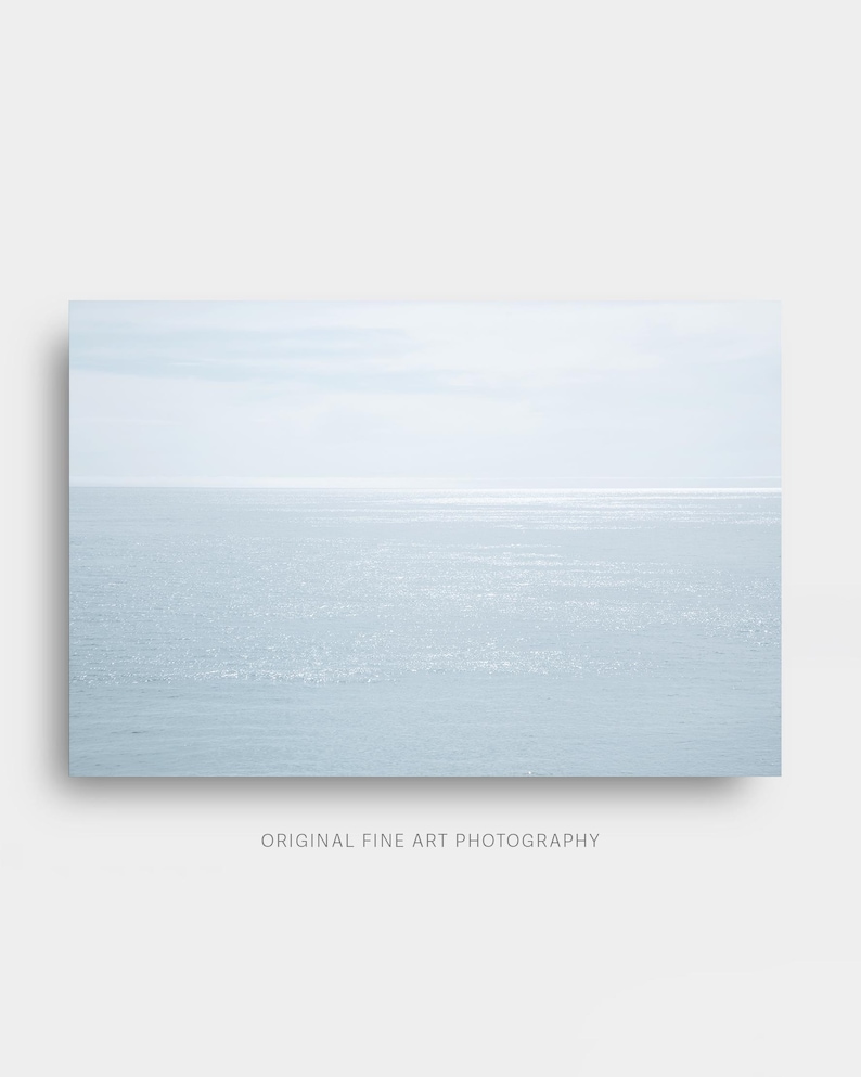 May include: A serene seascape photograph featuring a pale blue ocean meeting a light sky. The horizon line is visible, with subtle reflections of sunlight on the water's surface. The text "ORIGINAL FINE ART PHOTOGRAPHY" is at the bottom.