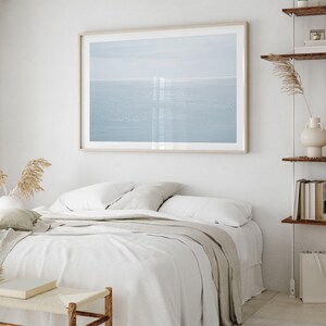 May include: A framed print of a seascape hangs above a bed with white linens and a light gray throw. A small wooden bench sits at the foot of the bed. The room has a minimalist, neutral color palette with wooden accents.