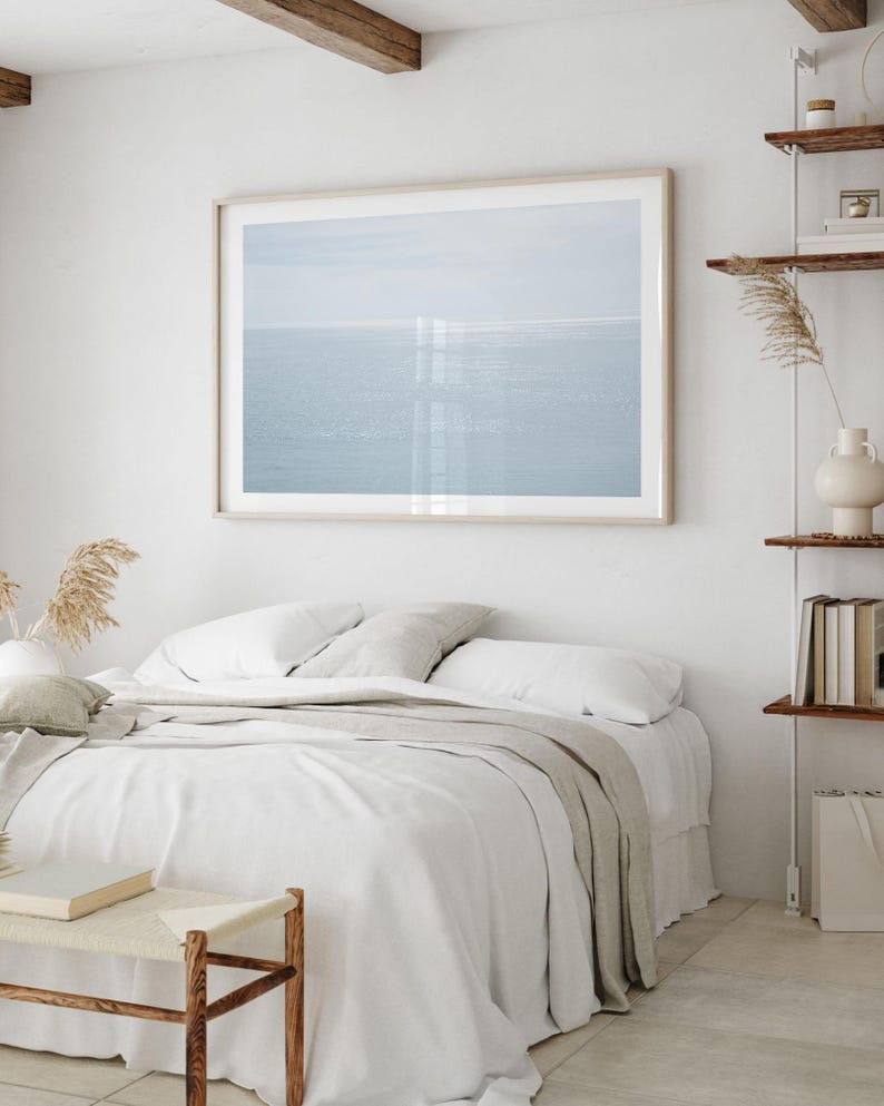 May include: A framed print of a seascape hangs above a bed with white linens and a light gray throw. A small wooden bench sits at the foot of the bed. The room has a minimalist, neutral color palette with wooden accents.