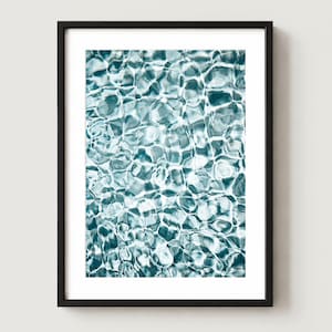 May include: A framed wall art print featuring a high-contrast photograph of rippling water. The image displays shades of deep teal, blue, and bright white light reflections, captured in a repeating pattern of distorted, organic shapes within a black frame.