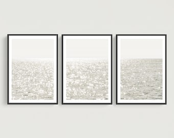 May include: A triptych of three vertical wall art prints, each framed in a thin black border. The prints display a monochromatic, high-contrast photograph of a sunlit ocean surface with bright white light reflections on the rippling water.