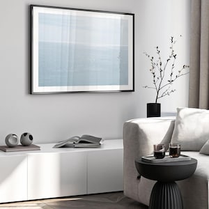 May include: A framed print of a seascape hangs on a white wall. The artwork features a soft blue and white color palette. A black side table holds a tray with two glasses. A black vase with a plant sits nearby.
