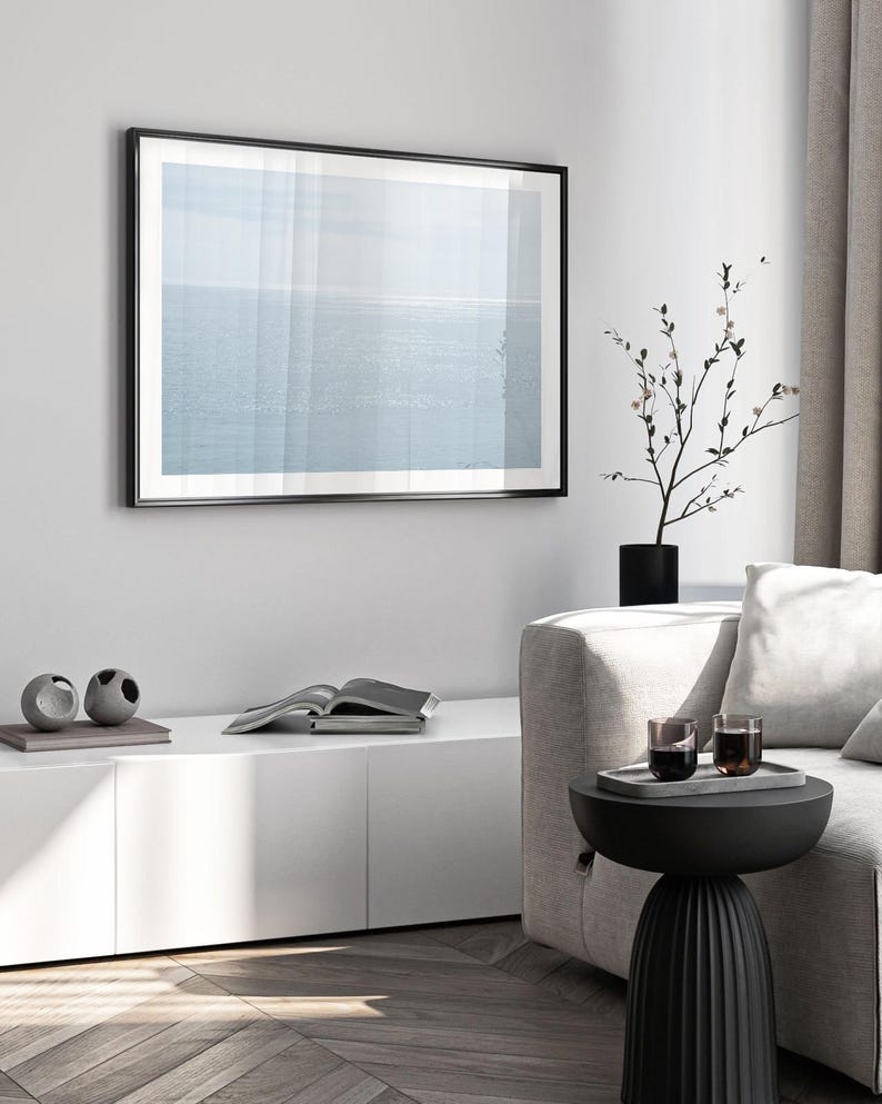 May include: A framed print of a seascape hangs on a white wall. The artwork features a soft blue and white color palette. A black side table holds a tray with two glasses. A black vase with a plant sits nearby.