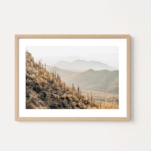 May include: A framed landscape print featuring a desert hillside covered in tall saguaro cacti. The scene displays a rocky slope in the foreground with layers of hazy, muted blue and tan mountains receding into the distance under a bright sky.
