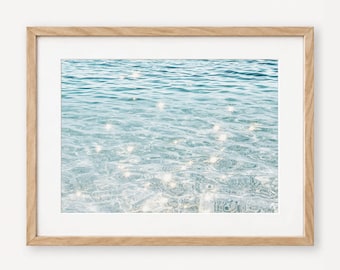 May include: A framed wall art print featuring a photograph of clear, rippling blue water with bright sunlight reflecting off the surface, creating numerous white star-like glints across the gentle waves in a light wood frame.