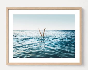 May include: A framed wall art print featuring a photograph of a pair of legs extending vertically out of the deep blue ocean water. The horizon line is visible in the distance under a clear sky, and the image is encased in a light wood frame.