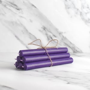 May include: A stack of purple taper candles tied together with twine. The candles are arranged in a pyramid shape on a white marble surface.