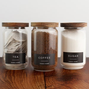 Puede incluir: Tres tarros de vidrio con tapas de madera y etiquetas negras. Las etiquetas dicen "Té", "Café" y "Azúcar". Los tarros están llenos de bolsitas de té, granos de café y azúcar.
