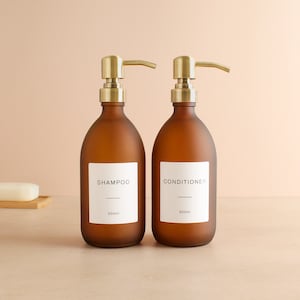 Puede incluir: Dos botellas de vidrio marrón esmerilado con bombas doradas y etiquetas blancas. La botella de la izquierda dice "Champú 500ml" y la botella de la derecha dice "Acondicionador 500ml".