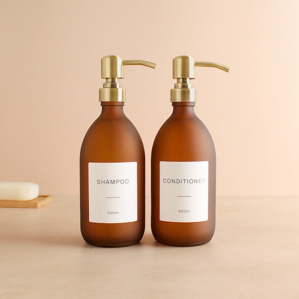 May include: Two frosted brown glass bottles with gold pumps and white labels. The bottle on the left says "Shampoo 500ml" and the bottle on the right says "Conditioner 500ml".