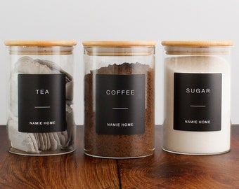 May include: Three clear glass jars with wooden lids, labeled "TEA", "COFFEE", and "SUGAR" in white text on black labels. The jars are filled with tea bags, coffee grounds, and sugar. The brand name "NAMIE HOME" is printed on the labels.
