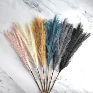 May include: A collection of faux pampas grass stems in various colors, including pink, cream, tan, teal, gray, and black. The stems have a feathery texture and are arranged against a white marble background. These decorative stems are suitable for home decor.