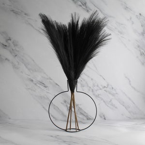 May include: A decorative black faux grass arrangement in a modern black metal vase. The vase has a circular design with three brown stems supporting the grass. The background is a white marble pattern.