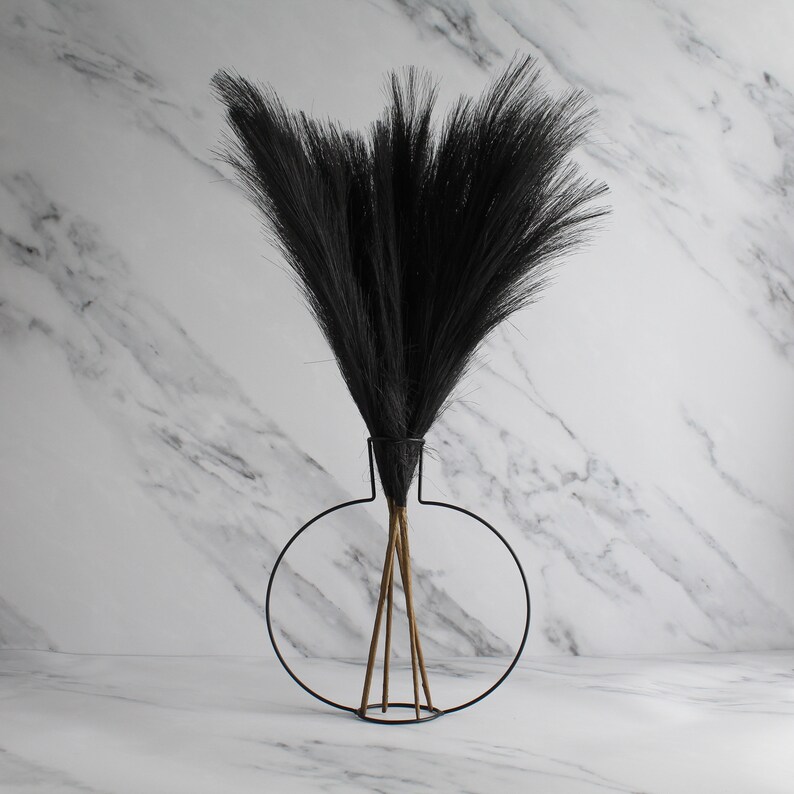 May include: A decorative black faux grass arrangement in a modern black metal vase. The vase has a circular design with three brown stems supporting the grass. The background is a white marble pattern.