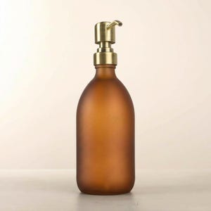 May include: A brown glass soap dispenser with a brass pump. The bottle has a matte finish and a rounded shape. The pump has a curved spout. The dispenser is designed for liquid soap or lotion.