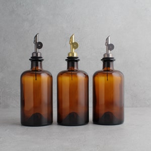 May include: Three amber glass bottles with different colored metal tops. The bottles have a narrow neck and a round body. The tops are black, gold, and silver.