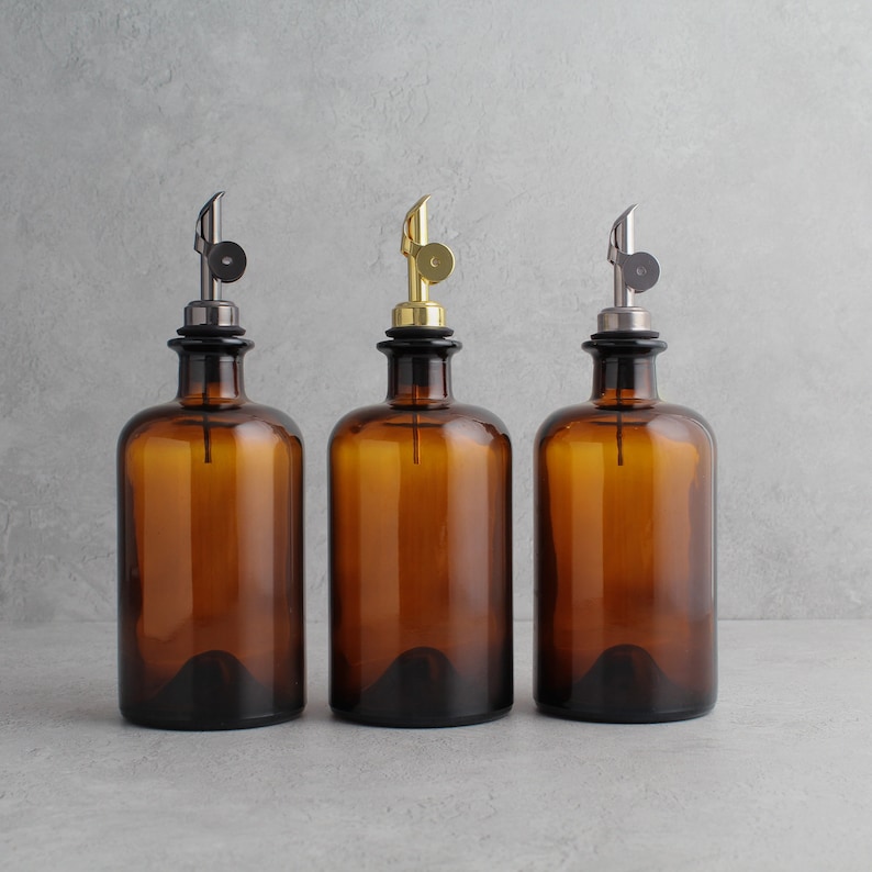 May include: Three amber glass bottles with different colored metal tops. The bottles have a narrow neck and a round body. The tops are black, gold, and silver.