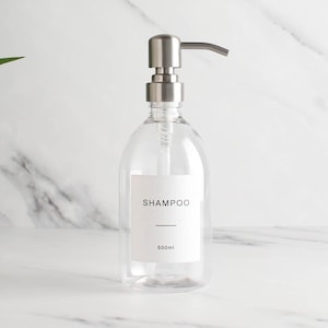Pode incluir: Frasco de champô de plástico transparente com um dispensador de bomba prateado. O frasco tem um rótulo branco com a palavra "CHAMPÔ" impressa em preto, juntamente com uma linha horizontal e a medida "500ml". O frasco está sobre um fundo de mármore branco.