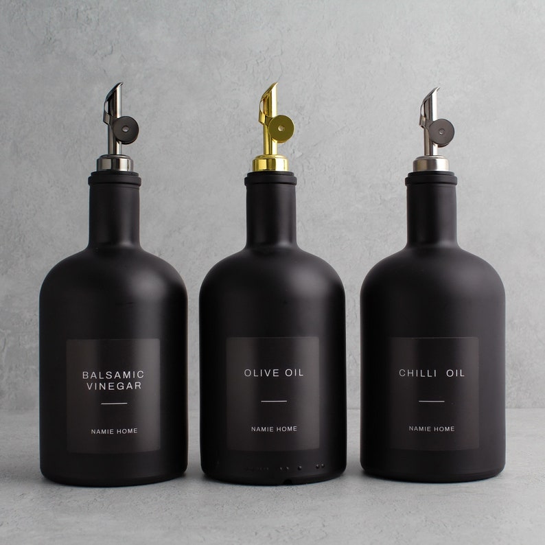 May include: Three black glass bottles with silver and gold pour spouts. The bottles are labeled "Balsamic Vinegar", "Olive Oil", and "Chilli Oil" with the text "NAMIE HOME" underneath each label.