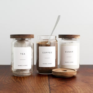 K&ouml;nnte beinhalten: Drei Glasbeh&auml;lter mit Holzdeckeln und Etiketten. Die Beh&auml;lter sind gef&uuml;llt mit Teebeuteln, Kaffeepulver und Zucker. Die Etiketten lauten "Tee", "Kaffee" und "Zucker". Die Beh&auml;lter stehen auf einer Holzoberfl&auml;che.