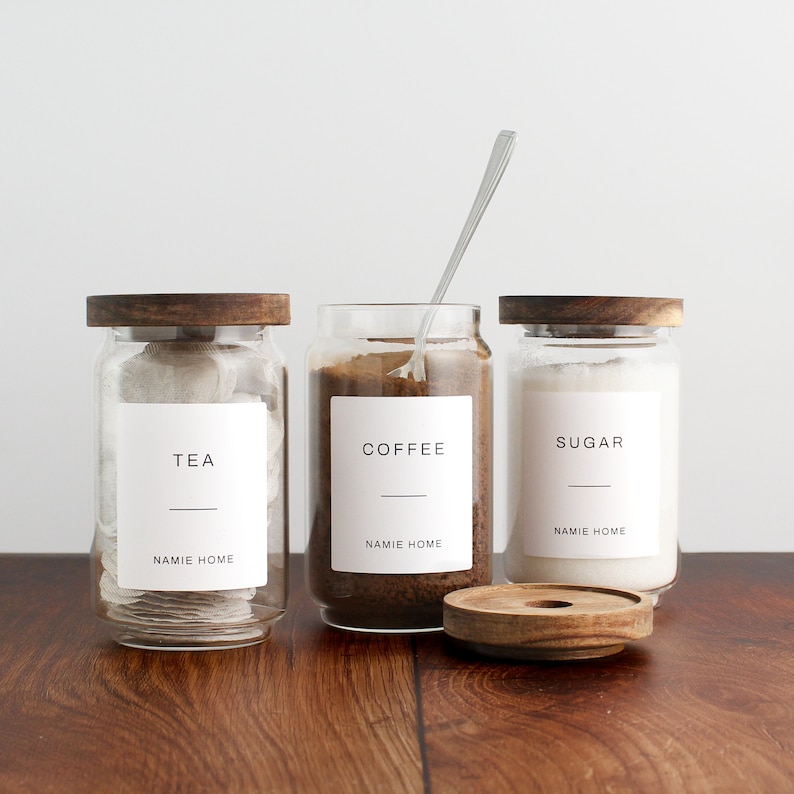 K&ouml;nnte beinhalten: Drei Glasbeh&auml;lter mit Holzdeckeln und Etiketten. Die Beh&auml;lter sind gef&uuml;llt mit Teebeuteln, Kaffeepulver und Zucker. Die Etiketten lauten "Tee", "Kaffee" und "Zucker". Die Beh&auml;lter stehen auf einer Holzoberfl&auml;che.